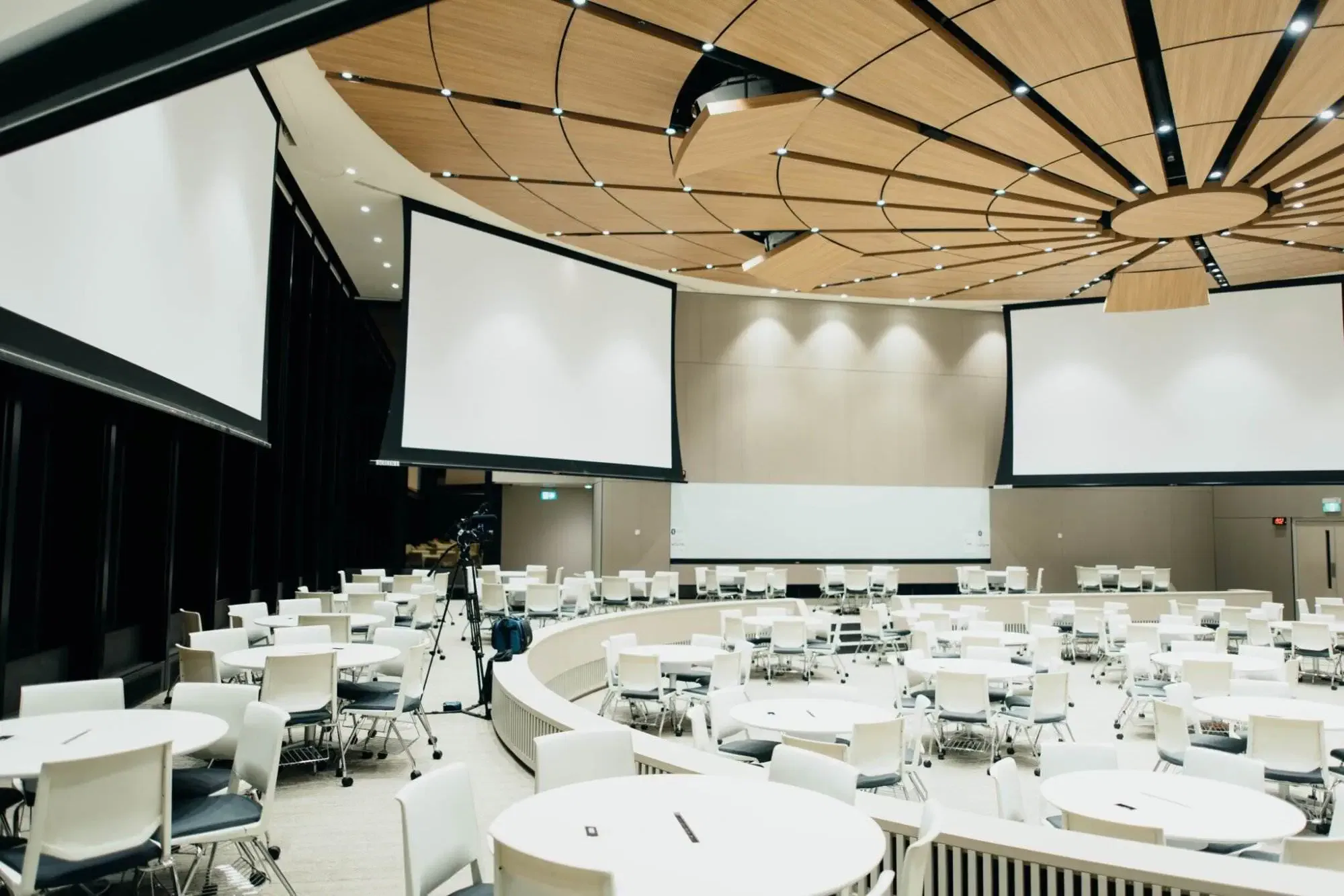 Een lichte, moderne conferentiezaal met ronde tafels en presentatieschermen, uitermate geschikt voor bijeenkomsten waarbij een tolk Hongaars de communicatie ondersteunt.